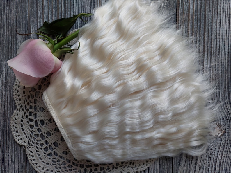 May include: A bundle of wavy, off-white hair extensions. A pink rose and a cream-colored lace doily are also in the image. The hair is styled in loose waves, and the overall aesthetic is soft and delicate.