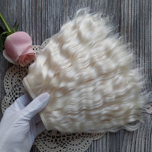 May include: A bundle of wavy, light blonde hair extensions. The hair is held by a gloved hand, with a pink rose and a lace doily in the background. The hair has a natural, soft texture and a light color.