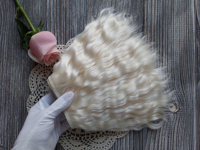 May include: A bundle of wavy, light blonde hair extensions. The hair is held by a gloved hand, with a pink rose and a lace doily in the background. The hair has a natural, soft texture and a light color.