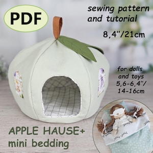 May include: A light green apple-shaped dollhouse with a brown handle and green leaf. The dollhouse has a large opening and two window cutouts with floral patterns. The text "APPLE HAUSE+ mini bedding" is visible. The dimensions are 21cm.