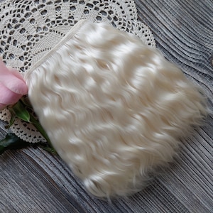May include: A bundle of wavy, off-white hair extensions. A pink rose and a cream-colored lace doily are also in the image. The hair extensions are likely made of natural fibers. The background is a gray wooden surface.
