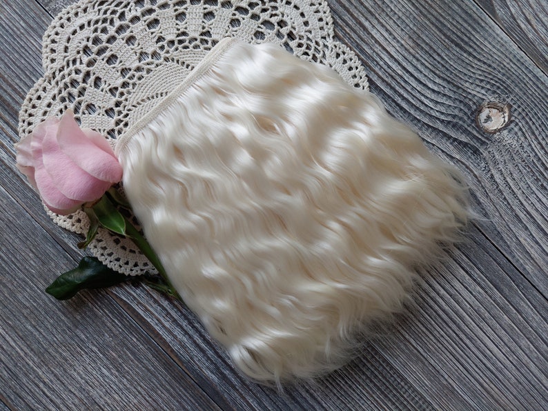 May include: A bundle of wavy, off-white hair extensions. A pink rose and a cream-colored lace doily are also in the image. The hair extensions are likely made of natural fibers. The background is a gray wooden surface.