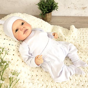 K&ouml;nnte beinhalten: Eine wei&szlig;e Baby-Puppe, die eine wei&szlig;e Strickm&uuml;tze, ein wei&szlig;es Langarmshirt und eine wei&szlig;e Hose tr&auml;gt. Die Puppe liegt auf einer wei&szlig;en Strickdecke.