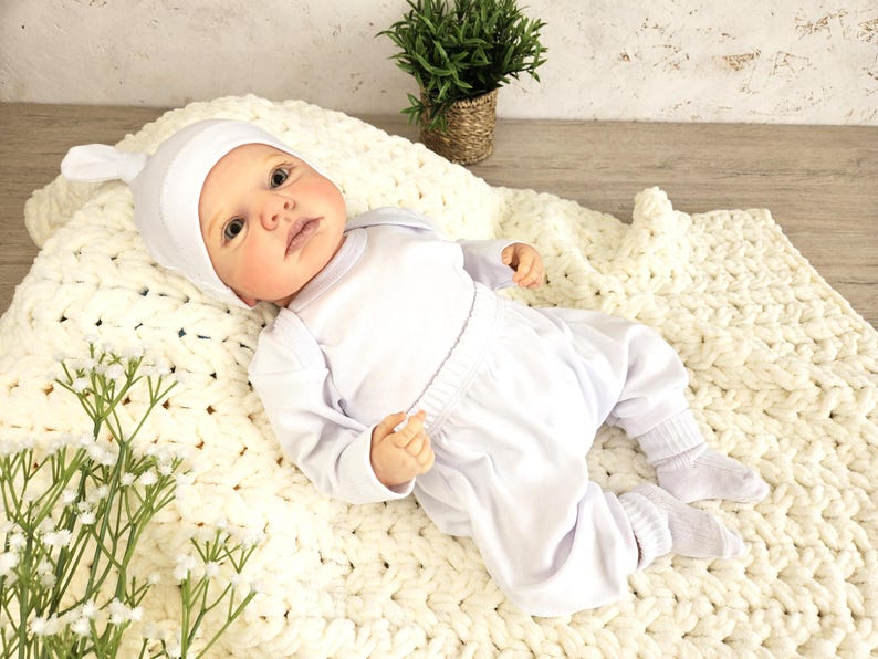 K&ouml;nnte beinhalten: Eine wei&szlig;e Baby-Puppe, die eine wei&szlig;e Strickm&uuml;tze, ein wei&szlig;es Langarmshirt und eine wei&szlig;e Hose tr&auml;gt. Die Puppe liegt auf einer wei&szlig;en Strickdecke.