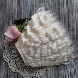 May include: A bundle of wavy, off-white doll hair rests on a cream-colored lace doily, next to a pink rose with green leaves. The background is a weathered, gray wooden surface.