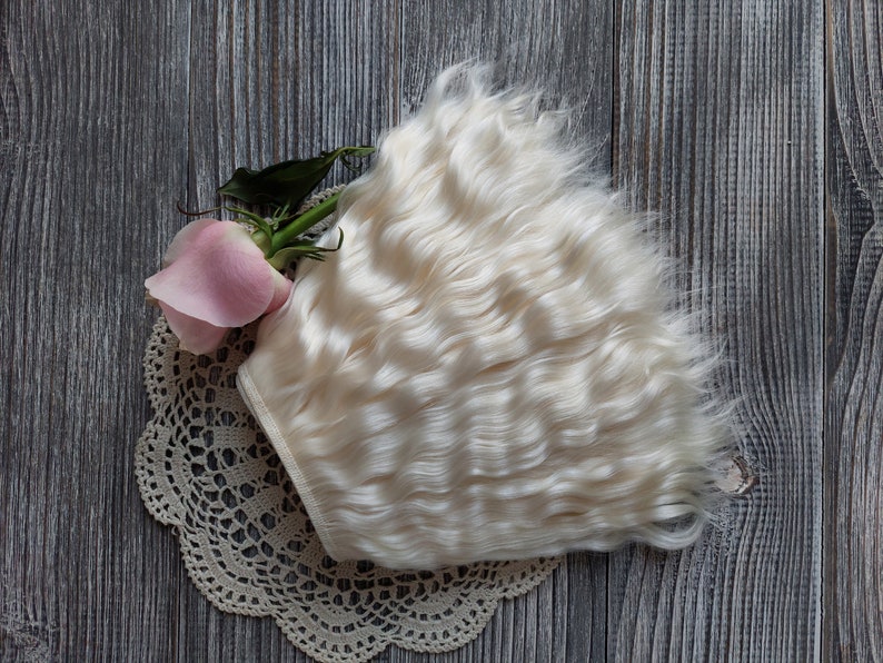 May include: A bundle of wavy, off-white doll hair rests on a cream-colored lace doily, next to a pink rose with green leaves. The background is a weathered, gray wooden surface.