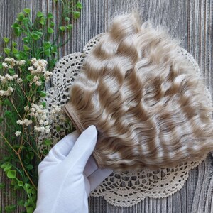 Puede incluir: Una trama de cabello ondulado rubio claro para el cabello de la mu&ntilde;eca. La trama est&aacute; sobre un encaje de encaje blanco y una mano enguantada de blanco la sostiene.