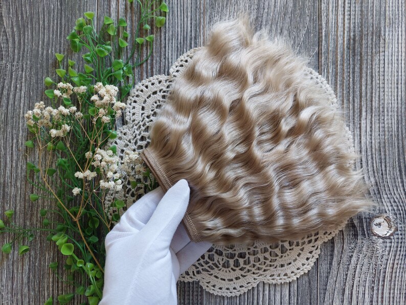 Puede incluir: Una trama de cabello ondulado rubio claro para el cabello de la mu&ntilde;eca. La trama est&aacute; sobre un encaje de encaje blanco y una mano enguantada de blanco la sostiene.