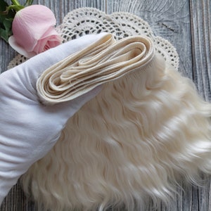May include: A bundle of light blonde doll hair, held by a gloved hand, with a wavy texture. A folded section of the hair is visible, along with a pink rose and a lace doily.
