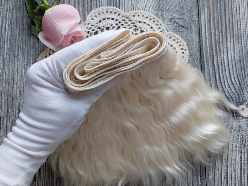 May include: A bundle of light blonde doll hair, held by a gloved hand, with a wavy texture. A folded section of the hair is visible, along with a pink rose and a lace doily.