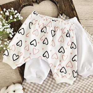 K&ouml;nnte beinhalten: Wei&szlig;e Baby-Shorts mit einem rosa und schwarzen Herz-Druck. Die Shorts haben einen wei&szlig;en elastischen Bund und sind aus weichem, bequemem Stoff.