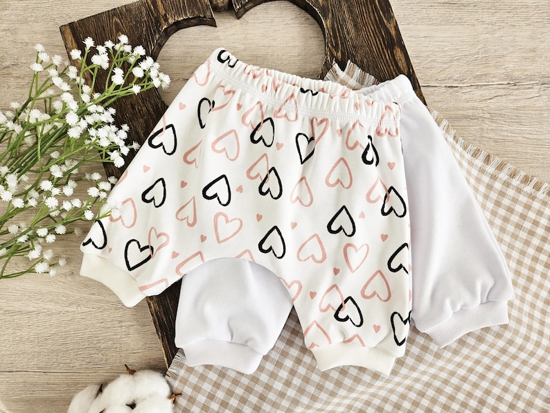 K&ouml;nnte beinhalten: Wei&szlig;e Baby-Shorts mit einem rosa und schwarzen Herz-Druck. Die Shorts haben einen wei&szlig;en elastischen Bund und sind aus weichem, bequemem Stoff.