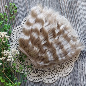 Puede incluir: Una trama de cabello ondulado rubio claro para hacer mu&ntilde;ecas. La trama est&aacute; sobre un encaje de encaje blanco sobre una superficie de madera.