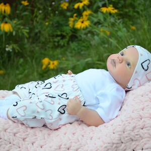 K&ouml;nnte beinhalten: Ein Baby tr&auml;gt einen wei&szlig;en Strampler mit einem rosa-schwarzen Herzmuster, eine wei&szlig;e M&uuml;tze mit rosa-schwarzen Herzen und wei&szlig;e Socken. Das Baby liegt auf einer rosa geh&auml;kelten Decke.