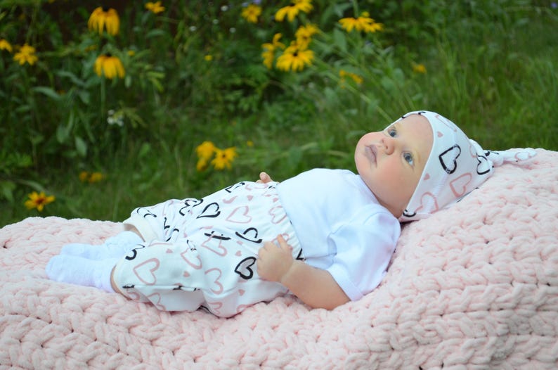 K&ouml;nnte beinhalten: Ein Baby tr&auml;gt einen wei&szlig;en Strampler mit einem rosa-schwarzen Herzmuster, eine wei&szlig;e M&uuml;tze mit rosa-schwarzen Herzen und wei&szlig;e Socken. Das Baby liegt auf einer rosa geh&auml;kelten Decke.