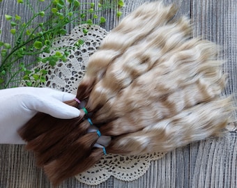 Peut inclure: Un ensemble de extensions de cheveux en deux nuances de blond, une claire et une plus foncée, attachées ensemble avec des élastiques colorés. Les extensions sont disposées en éventail sur un napperon en dentelle blanche.