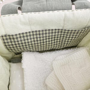 K&ouml;nnte beinhalten: Eine gesteppte Tragetasche mit einer Vichy-Tasche und grauen Griffen. Im Inneren befinden sich wei&szlig;e Decken und ein kleines Kissen. Die Tasche ist hellgr&uuml;n und wei&szlig;.