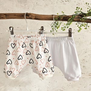 K&ouml;nnte beinhalten: Zwei Paar Baby-Bl&uuml;mchen, die an einem Kleiderb&uuml;gel h&auml;ngen. Ein Paar ist wei&szlig; und das andere ist wei&szlig; mit einem rosa und schwarzen Herzmuster.