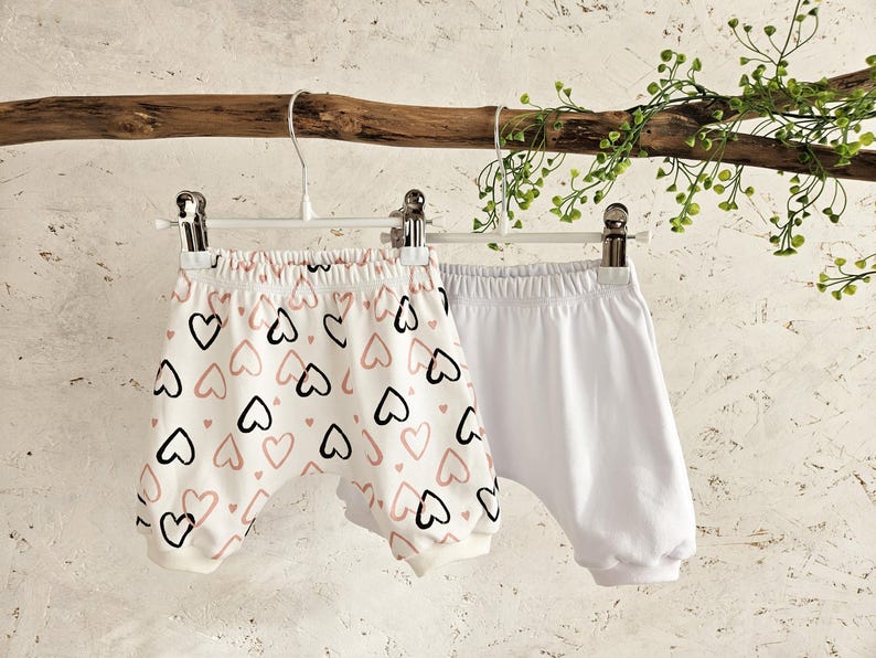 K&ouml;nnte beinhalten: Zwei Paar Baby-Bl&uuml;mchen, die an einem Kleiderb&uuml;gel h&auml;ngen. Ein Paar ist wei&szlig; und das andere ist wei&szlig; mit einem rosa und schwarzen Herzmuster.
