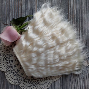 May include: A bundle of wavy, off-white hair extensions rests on a cream-colored lace doily, next to a pink rose with green leaves. The hair has a soft, natural texture. The background is a weathered, gray wooden surface.