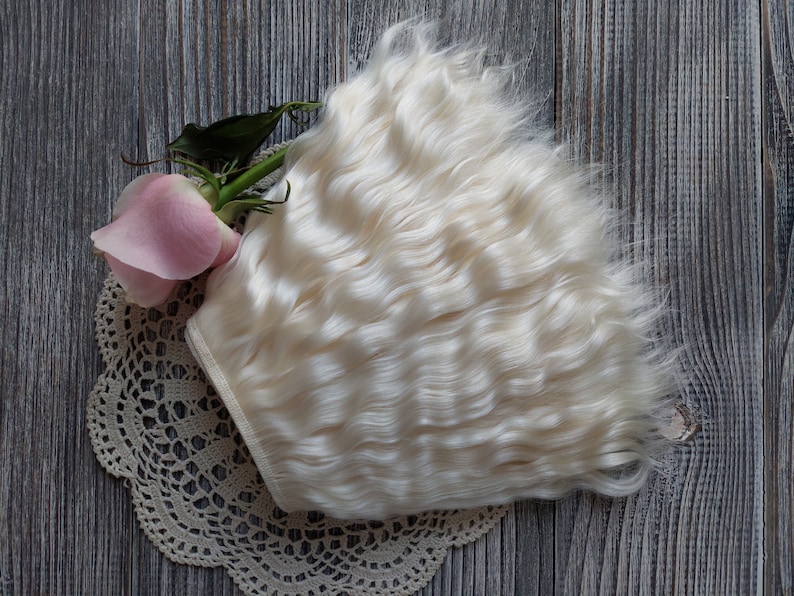 May include: A bundle of wavy, off-white hair extensions rests on a cream-colored lace doily, next to a pink rose with green leaves. The hair has a soft, natural texture. The background is a weathered, gray wooden surface.