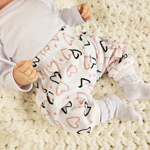 K&ouml;nnte beinhalten: Ein wei&szlig;er Baby-Strampler mit langen &Auml;rmeln und eine wei&szlig;e Hose mit einem pinkfarbenen und schwarzen Herzmuster.
