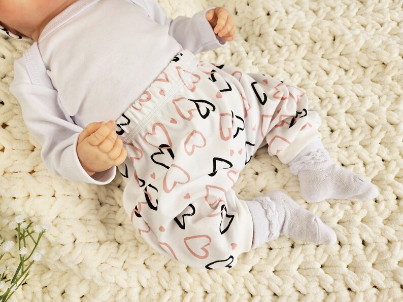 K&ouml;nnte beinhalten: Ein wei&szlig;er Baby-Strampler mit langen &Auml;rmeln und eine wei&szlig;e Hose mit einem pinkfarbenen und schwarzen Herzmuster.
