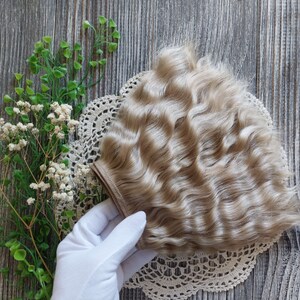 Puede incluir: Un paquete de extensiones de cabello humano rubio claro ondulado. El cabello est&aacute; dispuesto en forma de abanico y se sostiene en una mano enguantada.