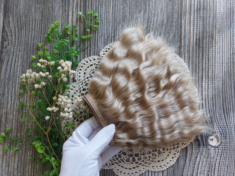 Puede incluir: Un paquete de extensiones de cabello humano rubio claro ondulado. El cabello est&aacute; dispuesto en forma de abanico y se sostiene en una mano enguantada.