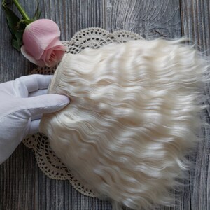 May include: A section of wavy, off-white hair extensions. The hair is held by a gloved hand, with a pink rose and lace doily in the background. The hair has a natural, textured appearance.