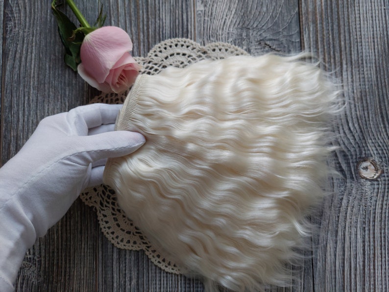 May include: A section of wavy, off-white hair extensions. The hair is held by a gloved hand, with a pink rose and lace doily in the background. The hair has a natural, textured appearance.