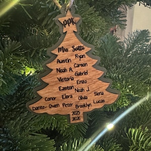 May include: A wooden Christmas tree ornament with a dark green outline and a light brown interior. The ornament has the initials "PPK" at the top and includes names and the years 2025 and 2026.