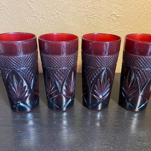 May include: Four ruby red drinking glasses with a detailed leaf and diamond pattern. The glasses have a slightly flared shape and are made of a dark red glass. The glasses are arranged in a row on a dark surface.