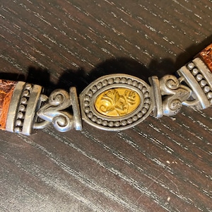 May include: A brown leather belt with a silver-toned buckle. The buckle has a gold-colored oval center with a floral design. The belt has silver-toned accents and a decorative design.