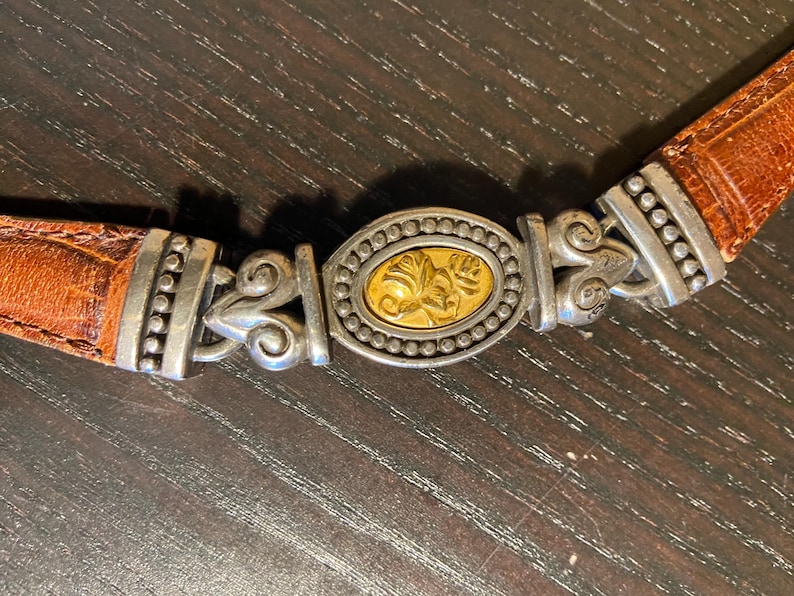 May include: A brown leather belt with a silver-toned buckle. The buckle has a gold-colored oval center with a floral design. The belt has silver-toned accents and a decorative design.