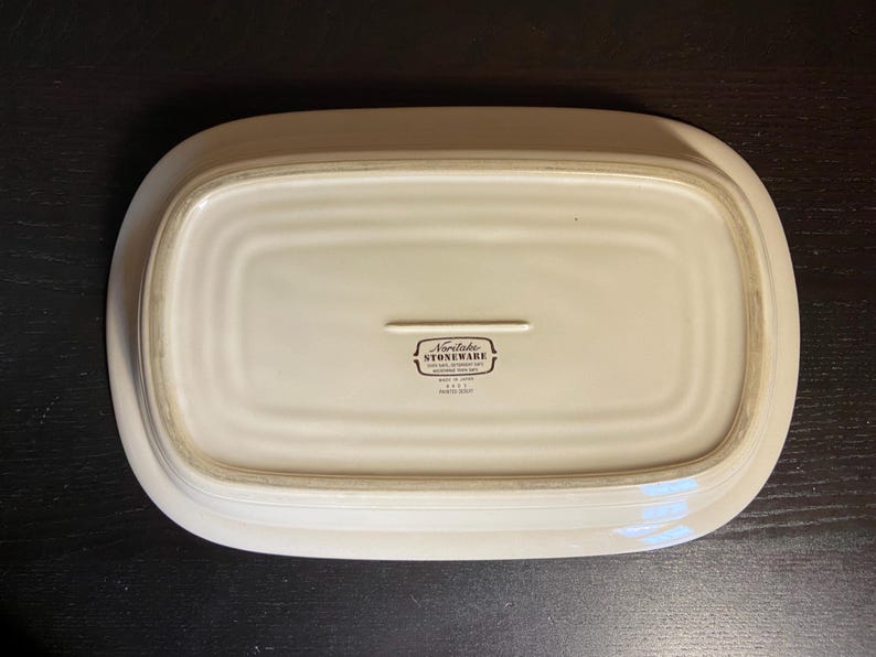 May include: Off-white rectangular stoneware serving platter. The underside of the platter is visible, with a recessed center and a raised border. The Noritake Stoneware logo is printed in black near the center.