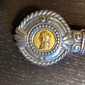 May include: A silver-toned belt buckle with a gold-colored center featuring a profile. The buckle has a rope-like border and is attached to a brown leather belt. The design is ornate and detailed.