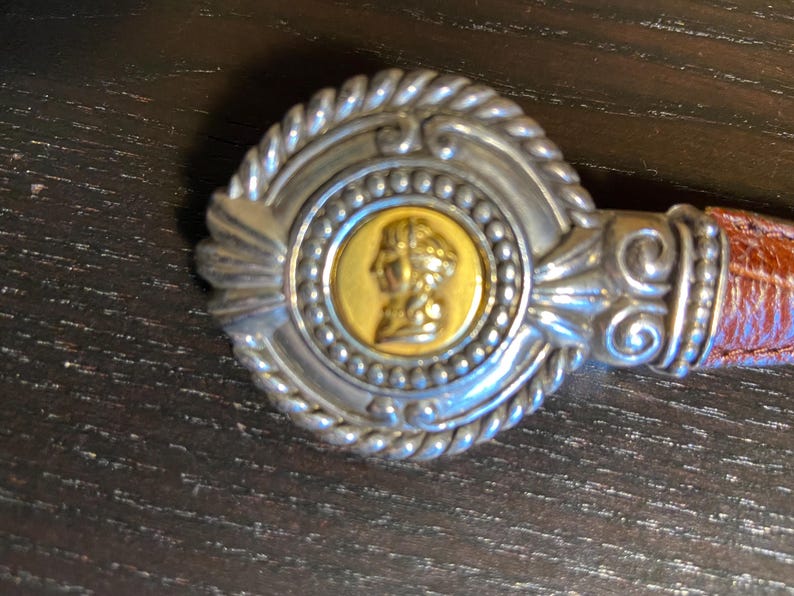 May include: A silver-toned belt buckle with a gold-colored center featuring a profile. The buckle has a rope-like border and is attached to a brown leather belt. The design is ornate and detailed.