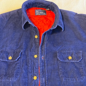 May include: A navy blue corduroy shirt with a red quilted lining. The shirt has two front pockets, gold buttons, and a collar. The label reads "The Men's Shop".