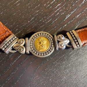 May include: A brown leather belt with a silver and gold buckle. The buckle features a gold coin with a profile of a woman's face, surrounded by silver beading. The belt is made of textured brown leather.