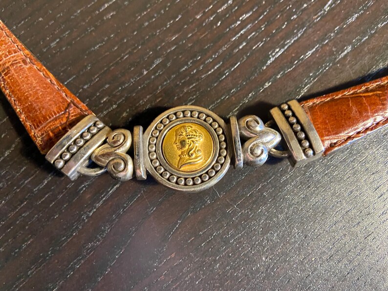 May include: A brown leather belt with a silver and gold buckle. The buckle features a gold coin with a profile of a woman's face, surrounded by silver beading. The belt is made of textured brown leather.