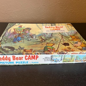 Puede incluir: Una caja de rompecabezas de imágenes vintage "Teddy Bear Camp". La caja presenta una colorida ilustración de osos de peluche acampando, con el texto "Teddy Bear Camp Picture Puzzle" y "TUCO". La caja es rectangular y tiene un borde blanco y azul.