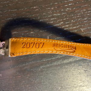 May include: Close-up of a tan leather belt strap with the number "20707" and the brand name "BRIGHTON" embossed. The strap is stitched with white thread and attached to a metal clasp.