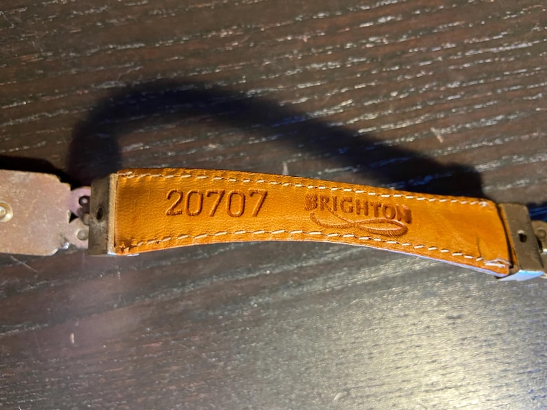 May include: Close-up of a tan leather belt strap with the number "20707" and the brand name "BRIGHTON" embossed. The strap is stitched with white thread and attached to a metal clasp.