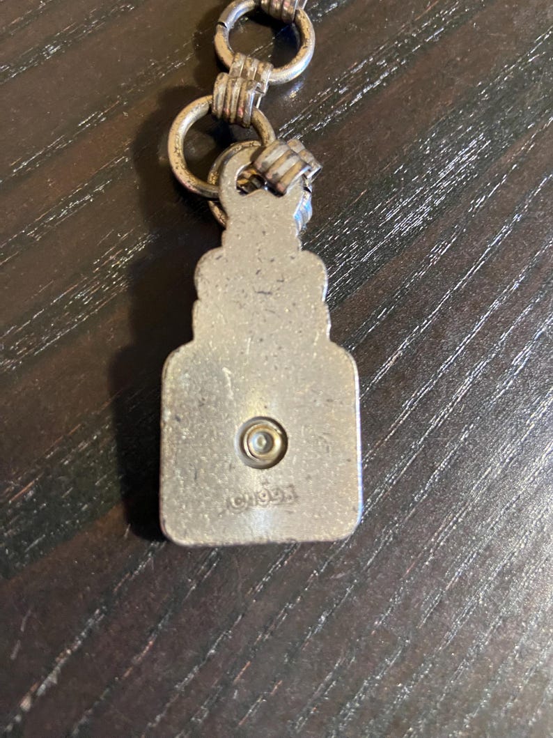 May include: A silver-toned metal pendant with a rectangular shape and a decorative top, connected to a chain with multiple rings. The pendant has a small circular detail and the text "CHOO93" is visible.
