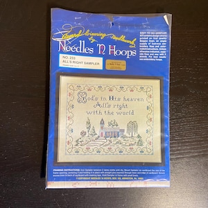 May include: A cross-stitch sampler kit by Needles 'n Hoops, featuring a framed design with the text "God's in His heaven, All's right with the world." The sampler includes a landscape scene with a church and trees, packaged in a blue plastic sleeve.