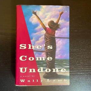 Puede incluir: Un libro de bolsillo titulado "She's Come Undone" de Wally Lamb. La portada presenta a una mujer con los brazos levantados, envuelta en una cuerda, contra un fondo de cielo y océano. El libro tiene un lomo rojo.