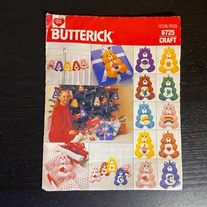 Puede incluir: Un patrón de manualidades Butterick vintage con Ositos Cariñositos. La portada es roja con el logotipo de Butterick y el número de patrón 6725. El patrón incluye imágenes de adornos de Ositos Cariñositos y una niña con un árbol de Navidad decorado con adornos de Ositos Cariñositos.