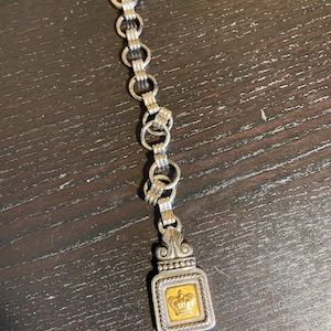 May include: A silver-toned chain with a rectangular pendant featuring a gold-colored square with a crown design. The chain is connected to a brown leather strap.