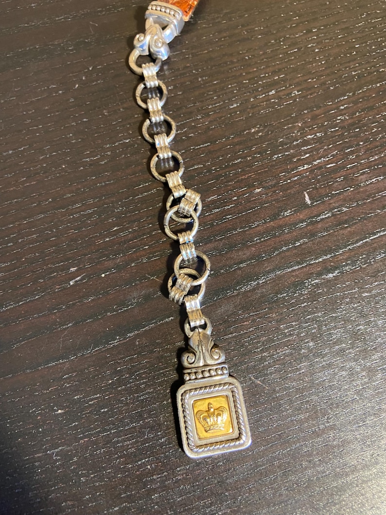 May include: A silver-toned chain with a rectangular pendant featuring a gold-colored square with a crown design. The chain is connected to a brown leather strap.